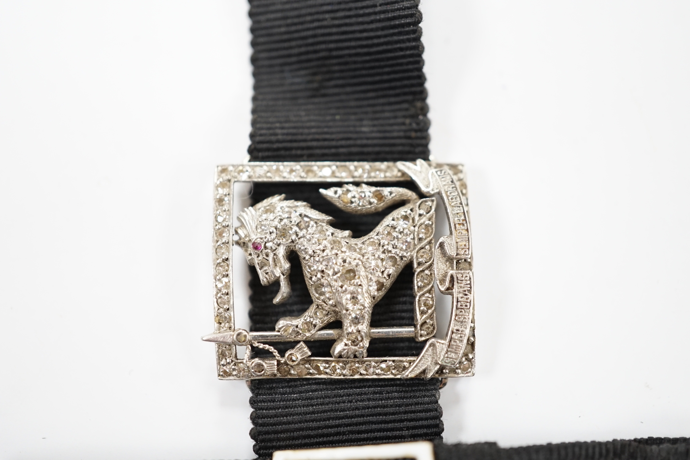 A black sash bracelet with diamond and enamel set Royal Fusiliers buckle, together with a similar sash with diamond set 'Charles' family crest, depicting a demi-rampant wolf, with motto.